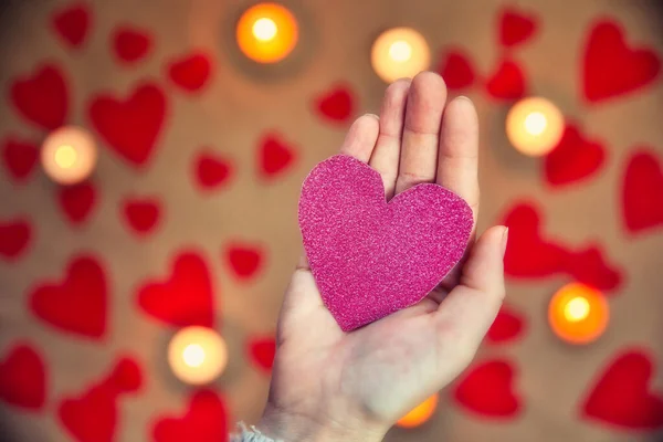 Mano femenina sosteniendo un corazón rosa brillante con vela romántica y fondo de corazones rojos para el día de San Valentín o concepto romántico —  Fotos de Stock