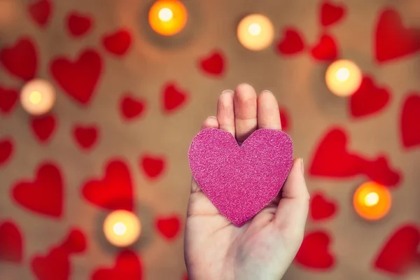 Mano femenina sosteniendo un corazón rosa brillante con vela romántica y fondo de corazones rojos para el día de San Valentín o concepto romántico —  Fotos de Stock