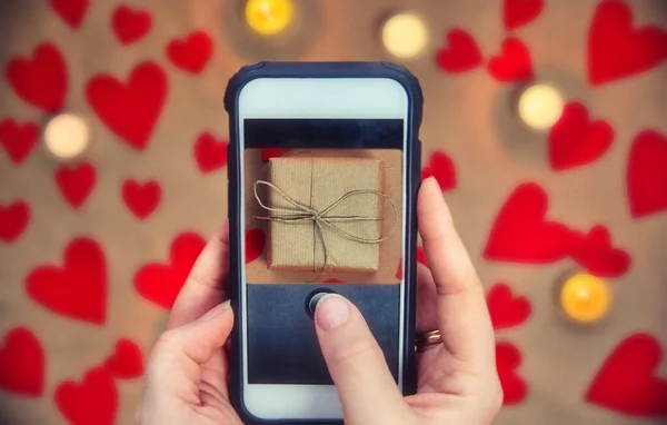 Kobieta trzyma telefon komórkowy robiąc zdjęcie pudełka na Walentynki, niewyraźne tło, romantyczny prezent w tle kartka okolicznościowa — Zdjęcie stockowe