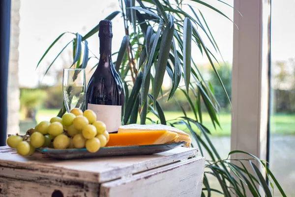 Cheese platter with grapes and wine on a table near a window with a cozy chair. Modern interior design — Stock Photo, Image