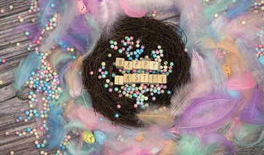 Colorful Easter eggs in bird nest surrounded with pastel colored feathers and wooden cubes with the text Happy Easter top view, modern background design, Easter holliday, Spring, concept