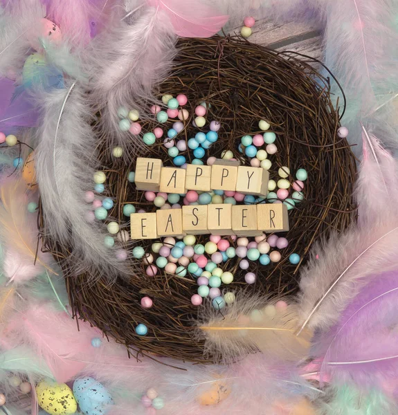 Colorful Easter eggs in bird nest surrounded with pastel colored feathers and wooden cubes with the text Happy Easter top view, modern background design, Easter holliday, Spring, concept — Zdjęcie stockowe