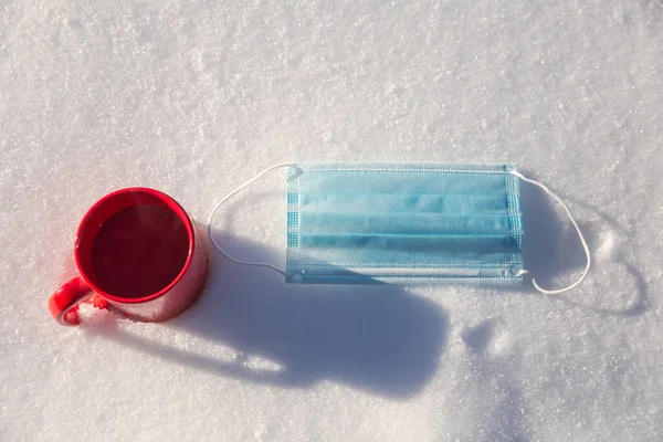 Red mug with hot coffee,tea or chocolate milk and protective blue medical mask for coronavirus Covid-19 pandemic. lies in the snow, Winter,virus,drink and health concept background — Stock Photo, Image
