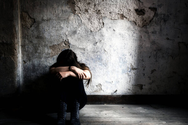 Depressed and lonely girl sitting alone in a spooky abandoned dirty old room on the wooden floor feeling miserable and anxiety cry over her life, depression and addiction issues concept with copy space shadows and dark place