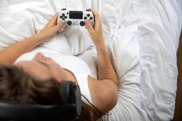 Woman playing video games with game controller while lying in bed, white sheets top view with copy space, Gaming,console concept concept