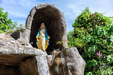 Statue of Virgin Mary in a rock cave chapel outside the St Anne Catholic Church in Baie Ste Anne, Praslin, Seychelles, with tropical vegetation around.