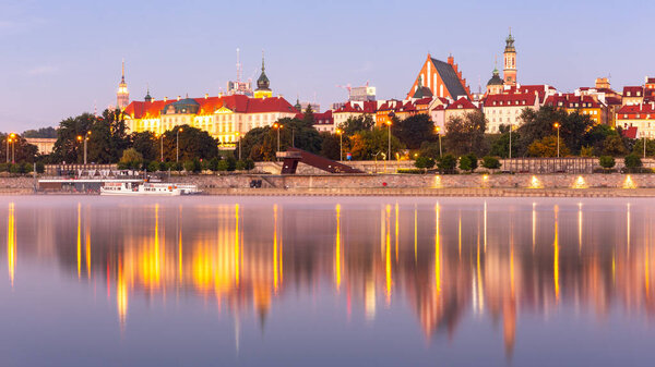 Освещенная панорама Старого города Варшавы с Королевским замком, собором и средневековыми зданиями, во время восхода солнца с отражениями в спокойной реке Висла, Польша.