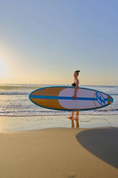 Jovem menina bonita em um maiô com uma prancha de surf ao amanhecer — Fotografia de Stock