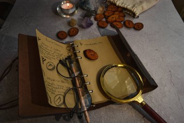 Masanın üzerinde rünler, bir kitap, bir büyüteç var.