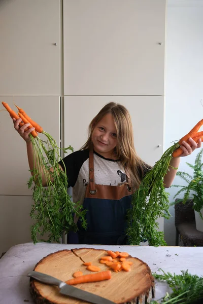 young girl in an apron in the kitchen with fresh carrots in her hands