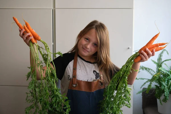 young girl in an apron in the kitchen with fresh carrots in her hands
