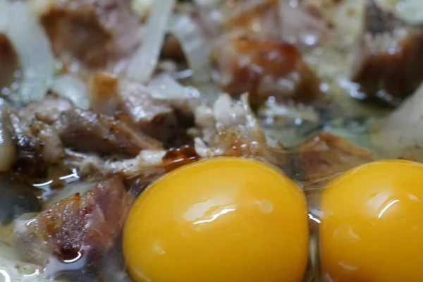 Las yemas de los huevos se rompen sobre los trozos de cerdo y cebolla que se cocinan en una sartén.. —  Fotos de Stock