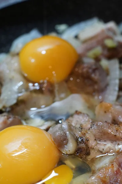 Las yemas de los huevos se rompen sobre los trozos de cerdo y cebolla que se cocinan en una sartén.. —  Fotos de Stock