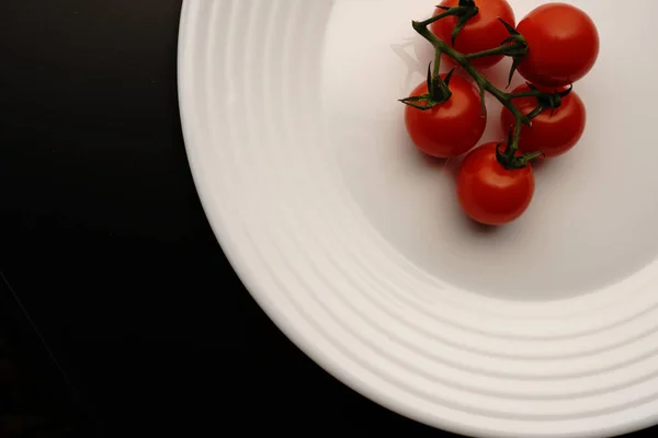 Os pequenos tomates vermelhos em um ramo verde estão em uma chapa branca em um contexto preto. Vista superior. — Fotografia de Stock