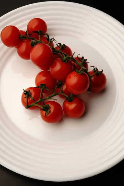 Os pequenos tomates vermelhos em um ramo verde estão em uma chapa branca em um contexto preto. Vista superior. — Fotografia de Stock