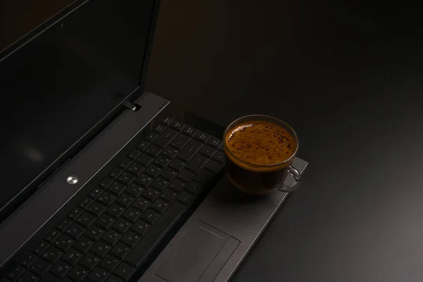 A laptop and a transparent cup of black coffee on a black background. Side view. Space for the text