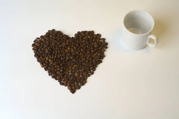 Un grande cuore fatto di chicchi di caffè è una tazza bianca su sfondo bianco. — Foto Stock