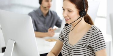 İnternette müşterilerle konuşurken kulaklık ve bilgisayar kullanan sıradan giyinmiş genç bir kadın. Bir grup operatör iş başında. Çağrı merkezi, iş konsepti