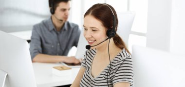 İnternette müşterilerle konuşurken kulaklık ve bilgisayar kullanan sıradan giyinmiş genç bir kadın. Bir grup operatör iş başında. Çağrı merkezi, iş konsepti