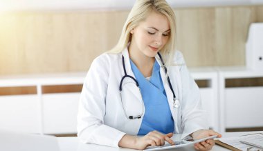 Kadın doktor güneşli klinikte oturuyor ve tablet bilgisayar kullanıyor. Veriler ve tıp ve sağlık hizmetlerindeki en iyi hizmet