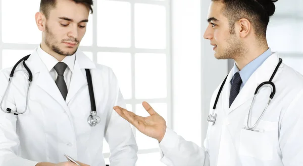 Médico árabe sentado en el escritorio de cristal con un colega caucásico en el consultorio médico o clínica. Equipo de médicos diversos, medicina y concepto de salud — Foto de Stock