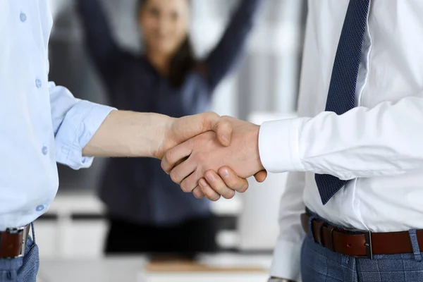 Dos hombres de negocios están estrechando las manos en la oficina, de cerca. Mujer de negocios feliz y emocionado se para con levantar las manos en el fondo. Concepto gente de negocios — Foto de Stock