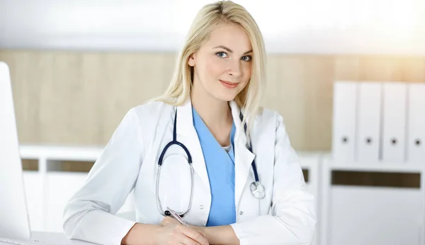 Ärztin bei der Arbeit, während sie am Schreibtisch in der sonnigen Klinik sitzt. Blonde fröhliche Ärztin füllt Krankenakte aus — Stockfoto