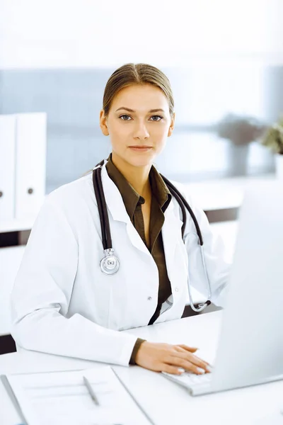 Woman-doctor typing on pc computer while sitting at the desk in hospital office. Physician at work