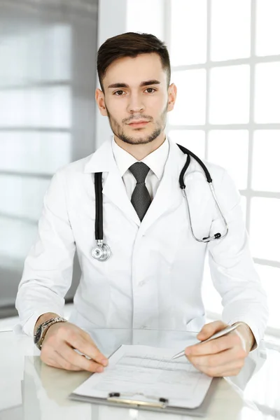 Lekarz wypełnia formularz historii leków siedząc przy szklanym biurku w klinice. Koncepcja medycyny i opieki zdrowotnej — Zdjęcie stockowe