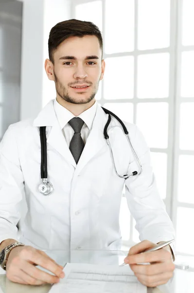 医者の男は診療所のガラスの机に座っている間に薬歴記録フォームを記入します。医学と医療の概念 — ストック写真