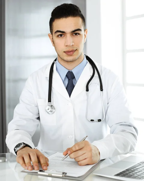 Arabische arts man gebruikt laptop computer tijdens het invullen van medicatie geschiedenis records formulier aan het glazen bureau in r kliniek. Geneesmiddelenconcept — Stockfoto