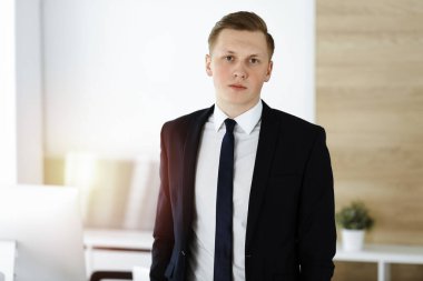 Neşeli iş adamı güneşli ofiste dikilip kameraya bakıyor. Genç girişimcinin vesikalığı