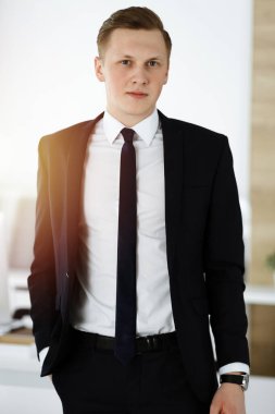 Neşeli iş adamı güneşli ofiste dikilip kameraya bakıyor. Genç girişimcinin vesikalığı