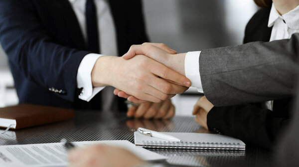Unknown businessman shaking hands with his colleague or partner above the glass desk in modern office, close-up. Business people group at meeting