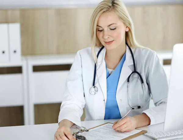 여자 - 의사가 병원이나 진료소에서 책상에 앉아 있는 동안에 말이죠. 의약 기록을 작성하고 있는 쾌활 한 의사 — 스톡 사진