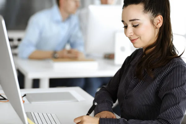 Mujer de negocios que utiliza la computadora en el lugar de trabajo en la oficina moderna. Secretaria morena o abogada mirando a la cámara y feliz sonriendo. Trabajar por placer y éxito —  Fotos de Stock