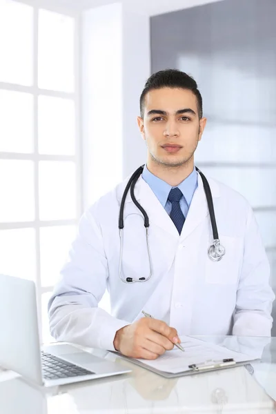 아랍 의사가 의료 사무실이나 진료소의 유리 책상에서 약물 기록을 작성하면서 노트북 컴퓨터를 사용하고 있다. 의학 과 건강 관리 개념 — 스톡 사진