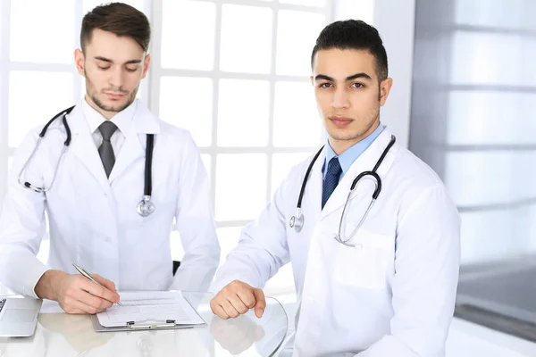 Médico árabe sentado en el escritorio de cristal con un colega caucásico en el consultorio médico o clínica. Equipo de médicos diversos, medicina y concepto de salud —  Fotos de Stock