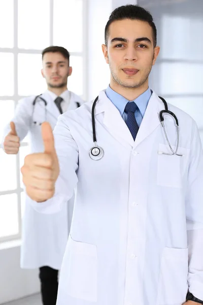 Arabische arts toont OK teken met duimen omhoog met Kaukasische collega in het medisch kantoor of kliniek. Diverse artsen team, beste behandeling, geneeskunde en gezondheidszorg concept — Stockfoto