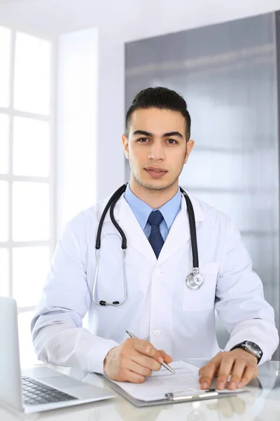 Hombre médico árabe que usa computadora portátil mientras llena el formulario de registros de historial de medicamentos en el mostrador de vidrio en la oficina médica o clínica. Concepto de medicina y salud —  Fotos de Stock