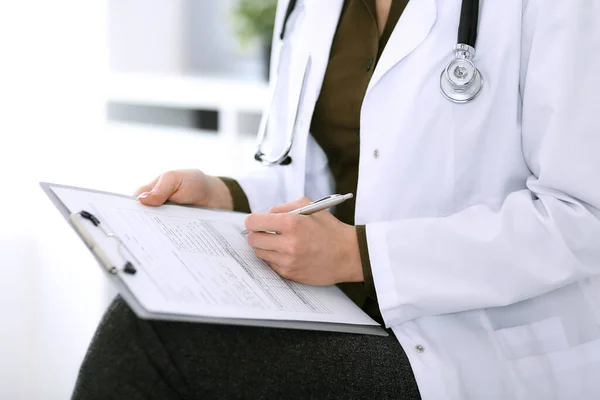 Doctora escribiendo algo en el portapapeles mientras está sentada en la silla, de cerca. Terapeuta en el trabajo llenando registros de historial de medicamentos. Concepto de medicina y salud — Foto de Stock