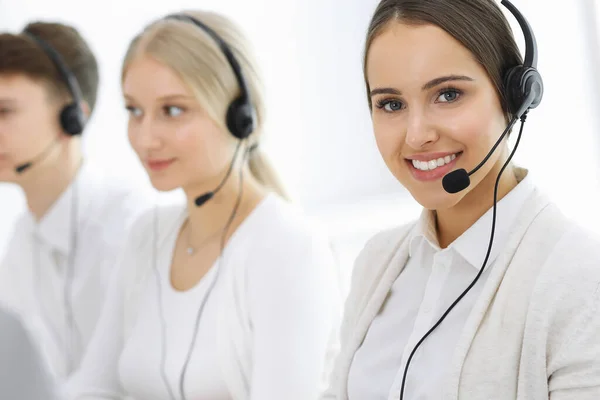 Call center. Group of operators at work. Focus on beautiful woman receptionist in headset at customer service white colored office. Business concept