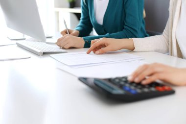 Muhasebeci mali durumu kontrol ediyor ya da vergi formları için hesap makinesi geliri ile sayıyor, eller yakın çekim. İş kadını ofisteki masada iş arkadaşıyla oturuyor ve çalışıyor. Denetim kavramı