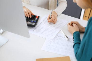 Muhasebeci mali durumu kontrol ediyor ya da vergi formları için hesap makinesi geliri ile sayıyor, eller yakın çekim. İş kadını ofisteki masada iş arkadaşıyla oturuyor ve çalışıyor. Denetim kavramı