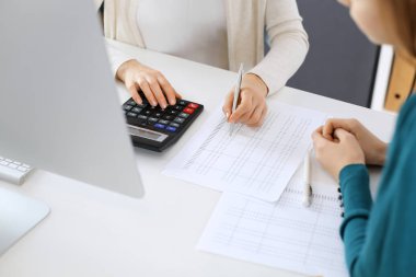 Muhasebeci mali durumu kontrol ediyor ya da vergi formları için hesap makinesi geliri ile sayıyor, eller yakın çekim. İş kadını ofisteki masada iş arkadaşıyla oturuyor ve çalışıyor. Denetim kavramı