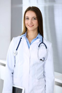 Doktor kadın mutlu ve neşeliyken arka planda hastane ofisinde dikiliyordu. Tıp ve sağlık hizmetleri kavramı