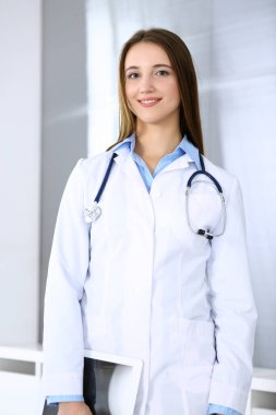 Doktor kadın mutlu ve neşeliyken arka planda hastane ofisinde dikiliyordu. Tıp ve sağlık hizmetleri kavramı