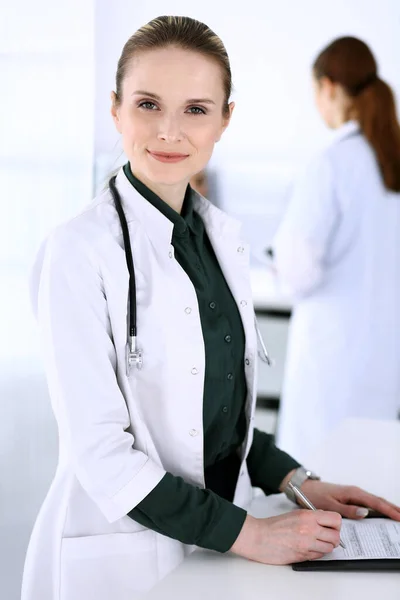 Doctora mujer en el trabajo con el paciente y colega en el fondo. Médico llenando documentos médicos o prescripción mientras está de pie en la recepción del hospital. Datos en medicina y asistencia sanitaria — Foto de Stock