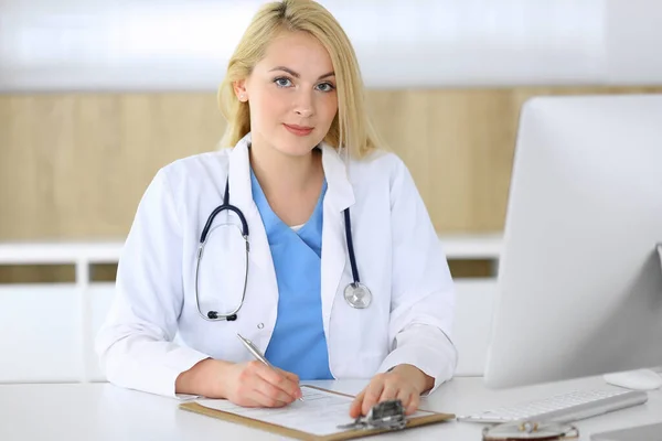 Médico mujer en el trabajo mientras está sentado en el escritorio en el hospital o clínica. Rubia médico alegre llenar el formulario de registros médicos. Datos y mejor servicio en medicina y salud — Foto de Stock