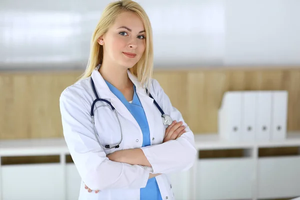 Doctor woman at work while standing with arms crossed in hospital or clinic. Blonde cheerful physician ready to help patients. Data and best service in medicine and healthcare